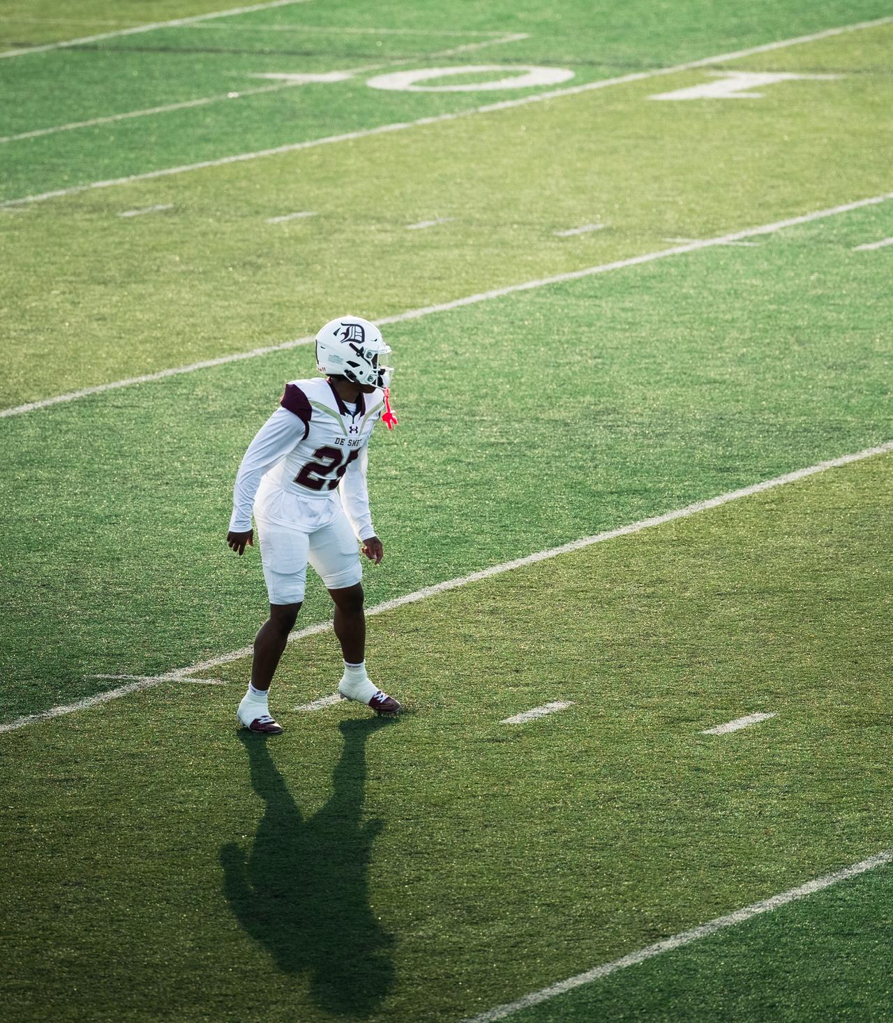 Lone defensive back in white and maroon standing on the field, casting a long shadow across the grass