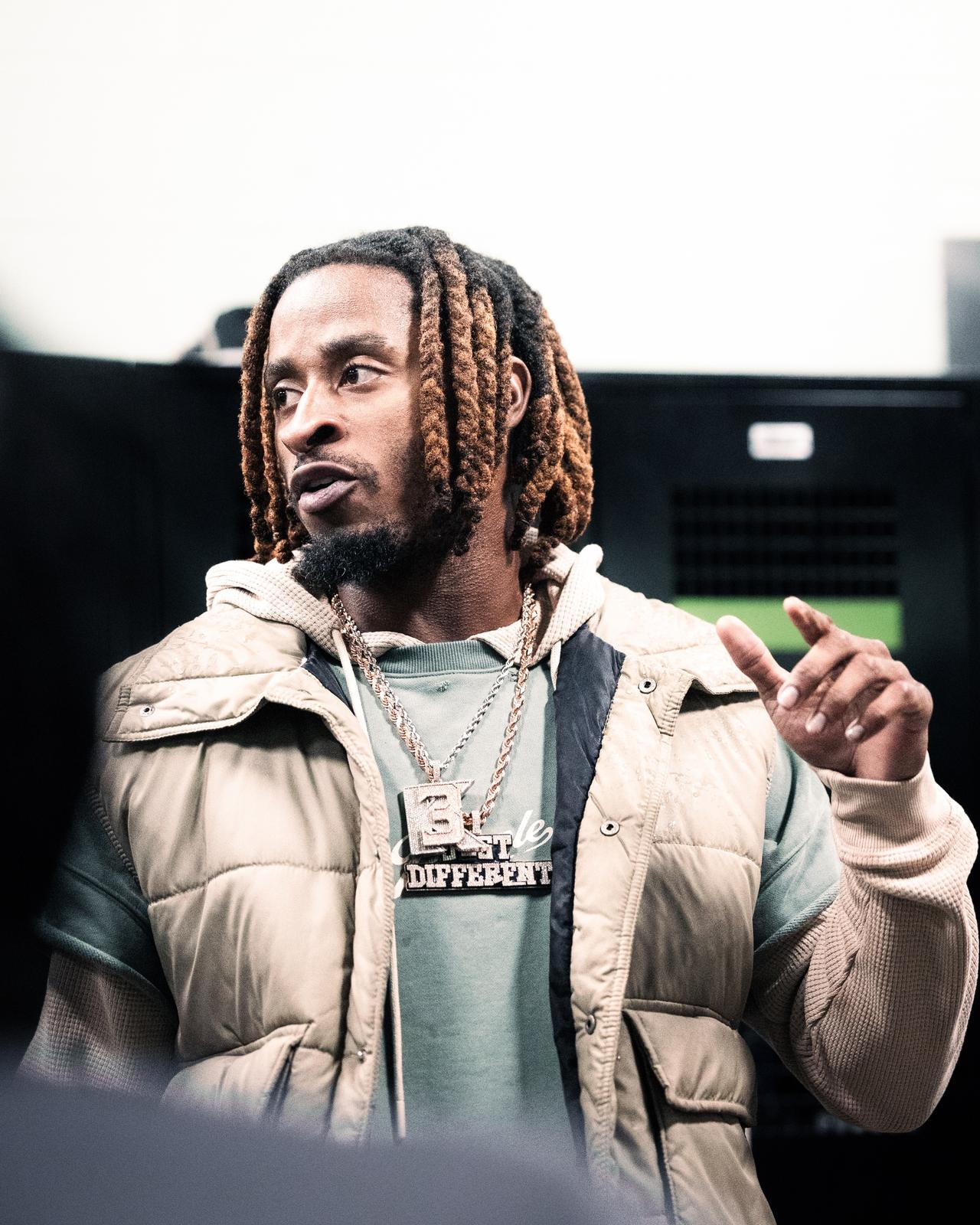 Man with copper-tipped locs in a tan puffer vest and chain, gesturing in a locker room