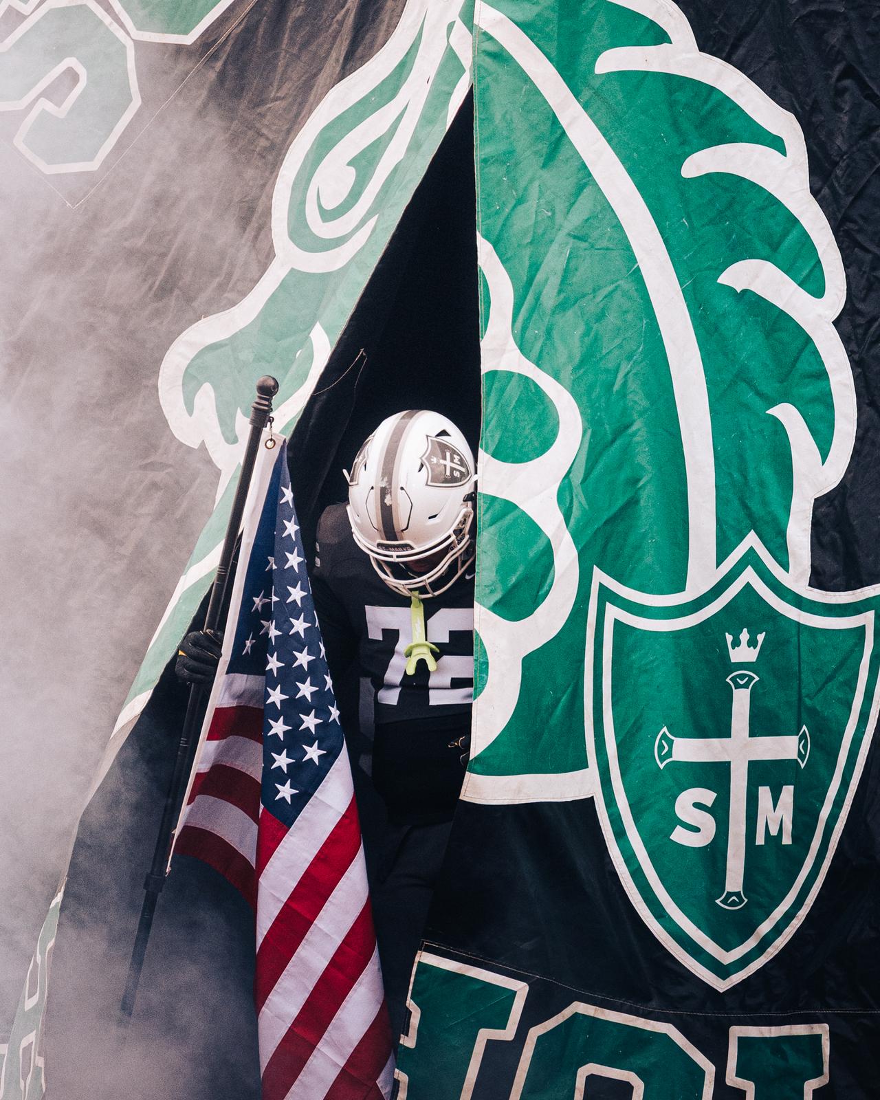 Player emerging through a green run-through banner holding an American flag, surrounded by smoke