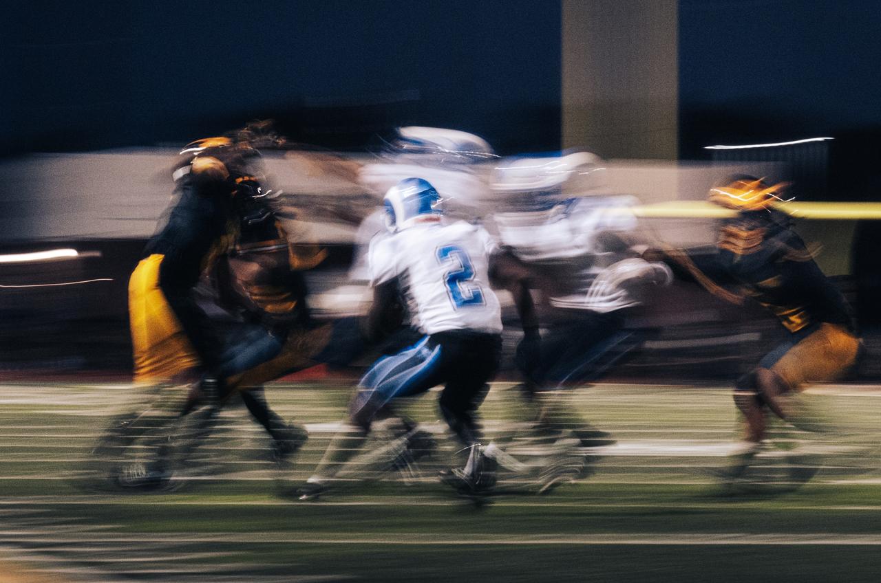 Motion-blurred panning shot of blue and white players against black and gold under stadium lights